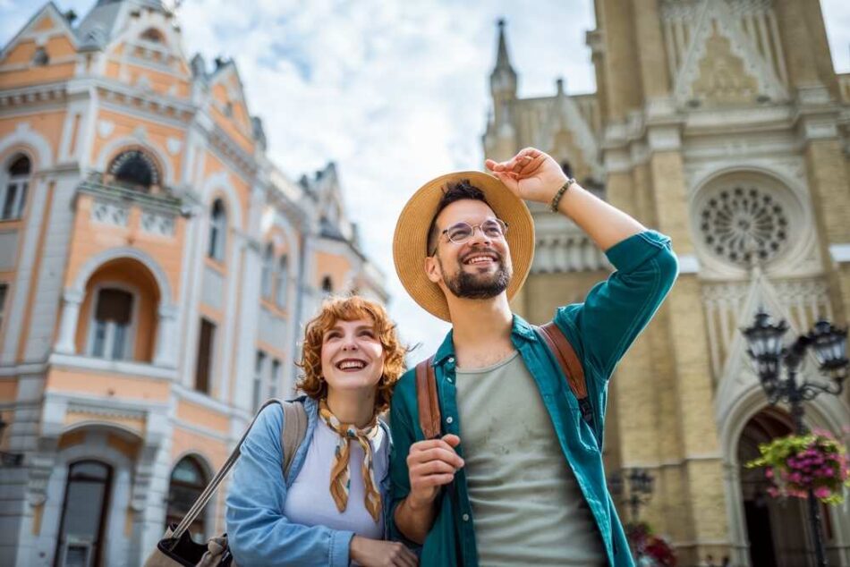 Mladý pár usmiatych turistov objavuje historické mesto s krásnou architektúrou. Muž nosí slamený klobúk a okuliare, žena má šatku okolo krku. Na pozadí sú historické budovy s detailnou fasádou a vežou.